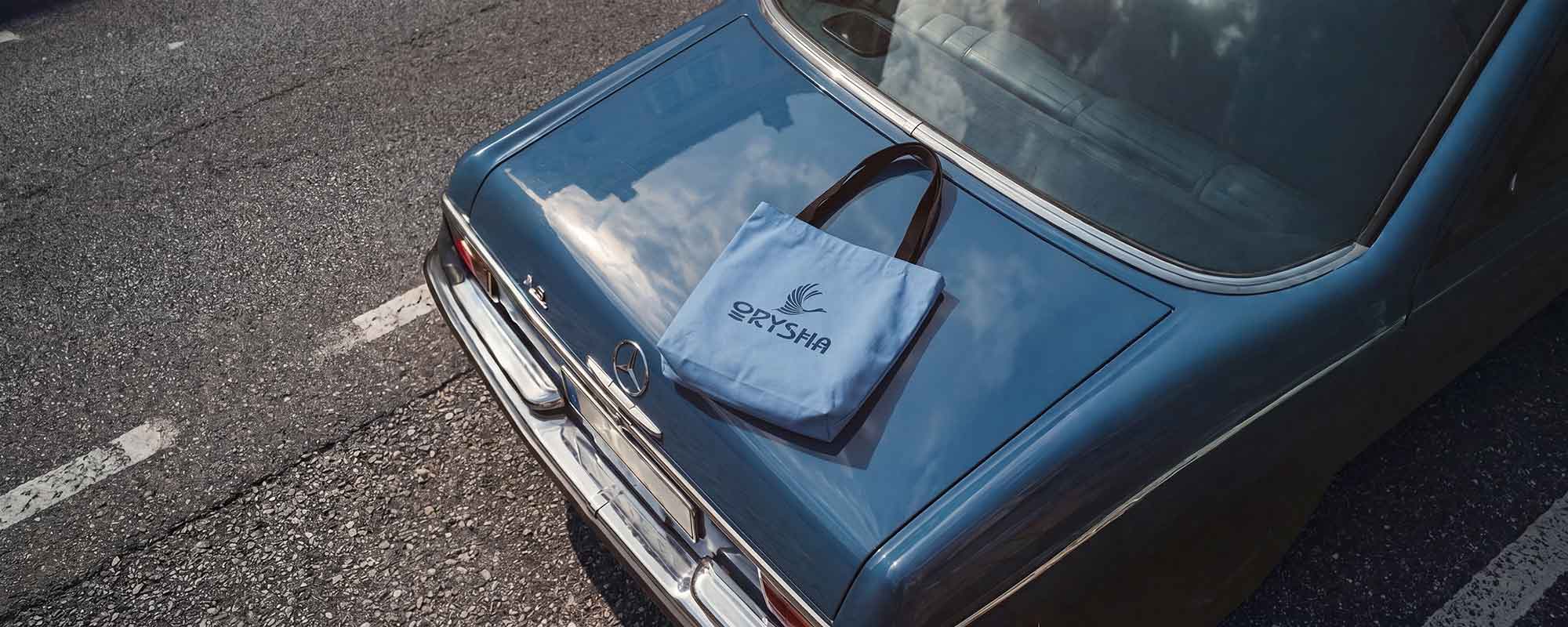 A light blue canvas tote bag with a dark brown handle and a white 'ORYSHA' logo printed on the front, resting on the trunk of a blue vintage car on an asphalt road.