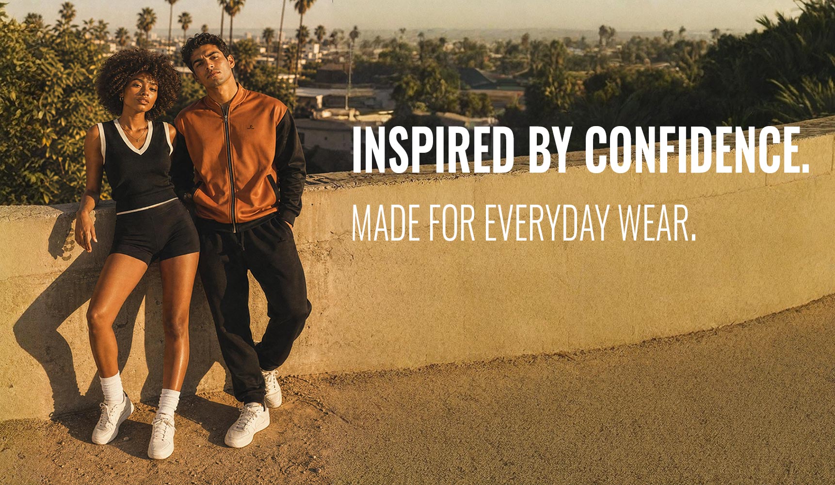 Two models posing outdoors in casual streetwear, with one wearing a black tank top and shorts and the other wearing an orange and black jacket with black pants. Large white text on the right reads 'INSPIRED BY CONFIDENCE.