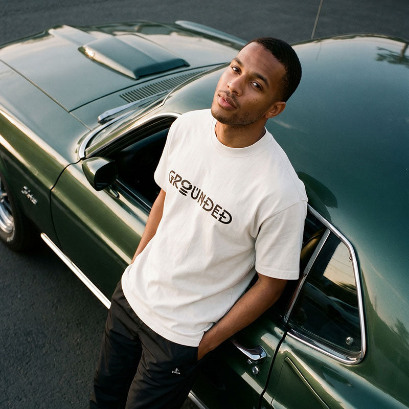 A man wearing a white t-shirt with 'GROUNDED' printed in black across the chest, leaning against a dark green classic car on an asphalt surface.