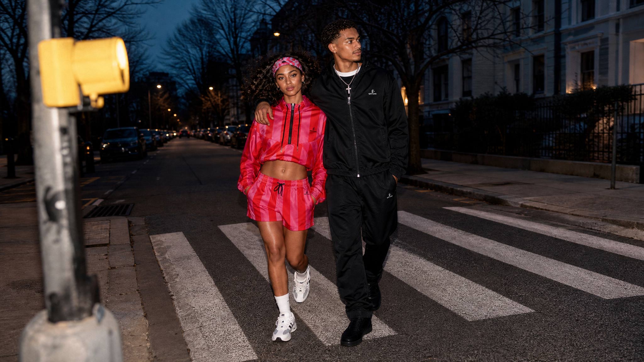 A man and a woman walk side-by-side across a city crosswalk at dusk. The woman is wearing a vibrant pink athletic tracksuit with a matching headband, while the man wears a black tracksuit.