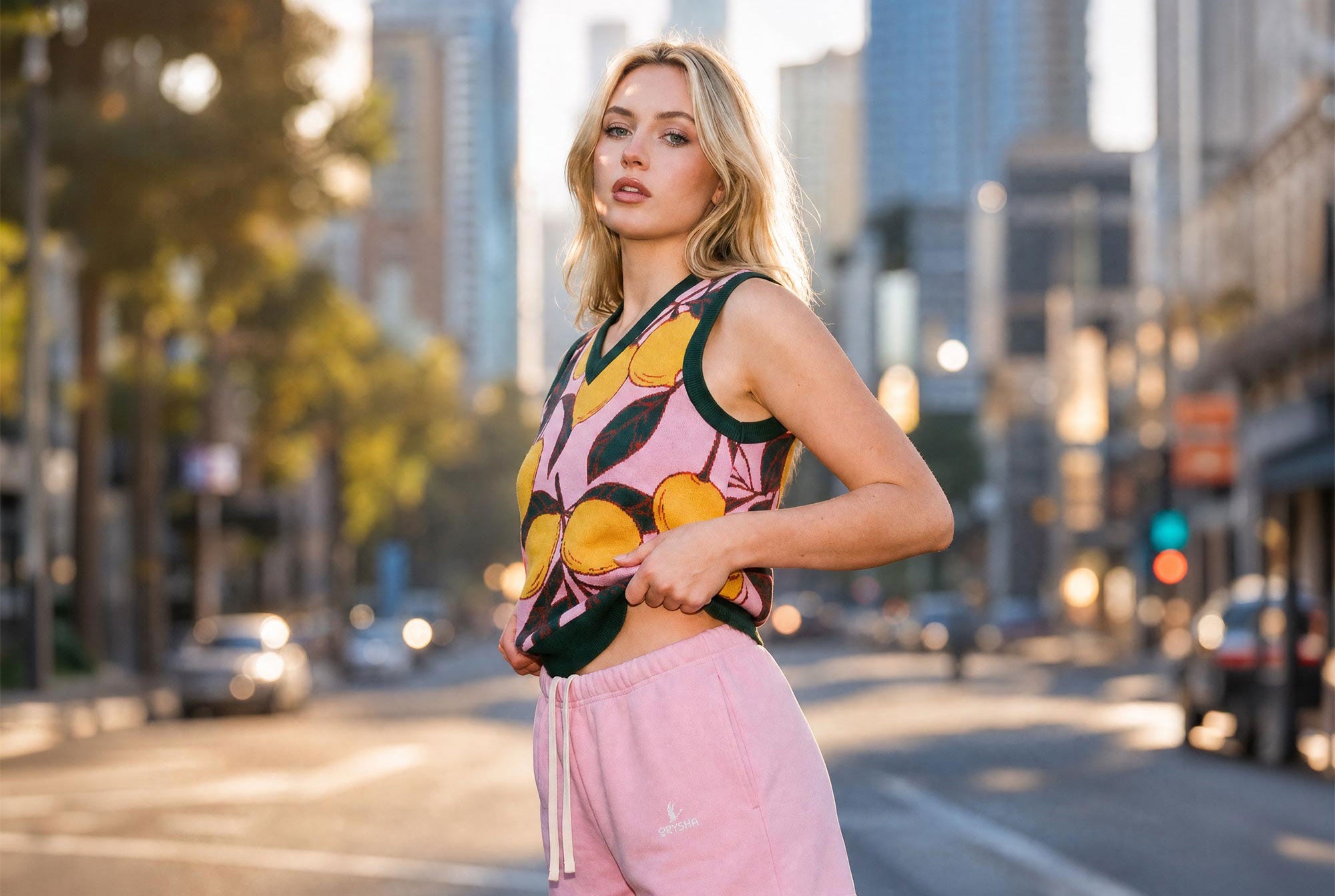 A woman stands on a city street at sunset wearing a sleeveless crop top with a pink, yellow, and black citrus pattern and light pink sweatpants.