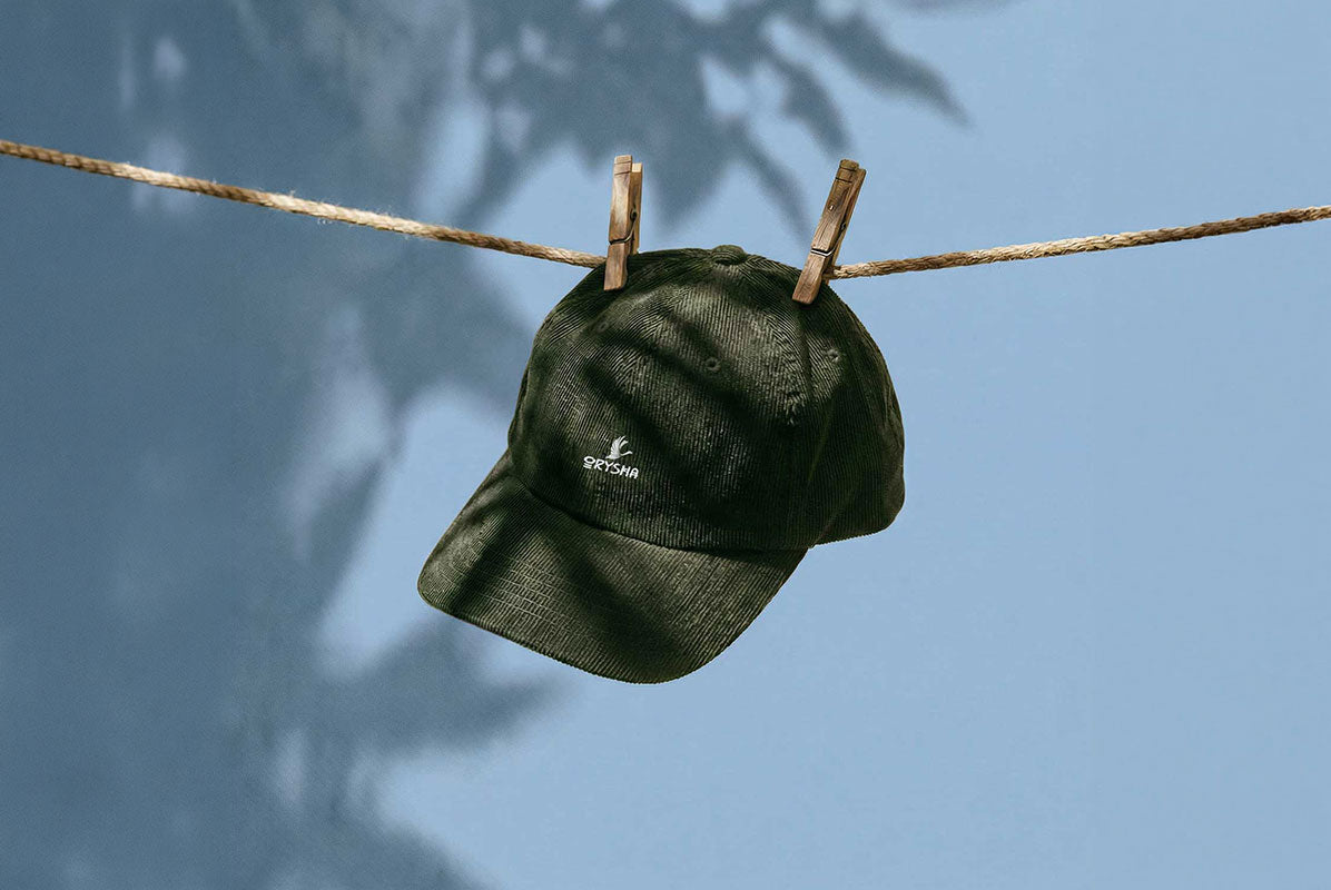 A dark olive green baseball cap with a small white embroidered logo, hanging from a rope with wooden clothespins against a blue sky with leaf shadows.
