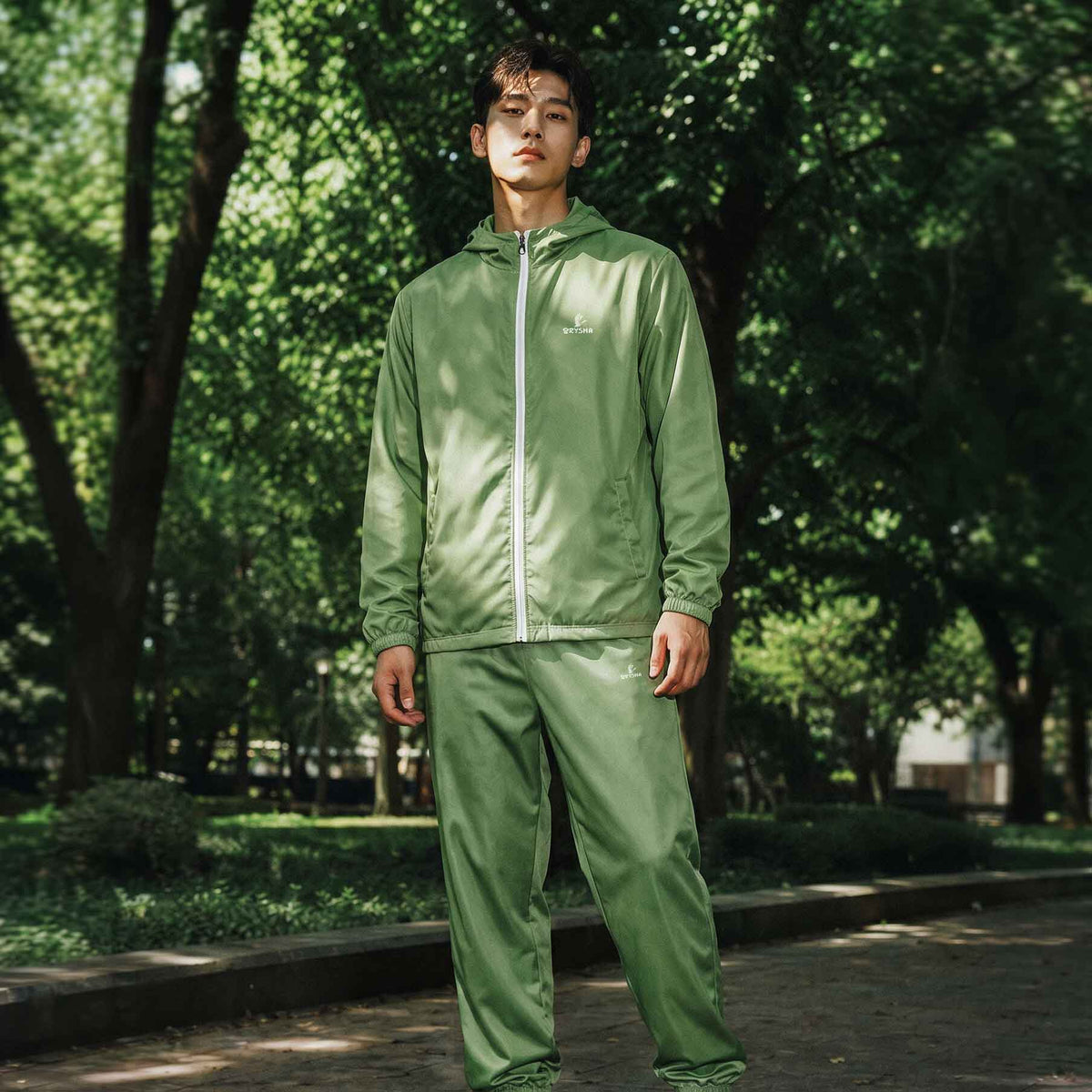 Person wearing a green track jacket and pants standing in a park with trees in the background