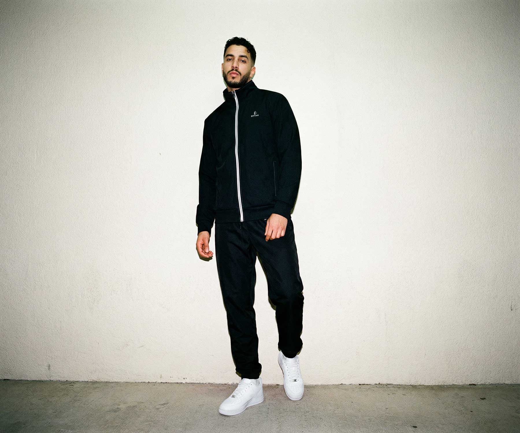 Man wearing a black tracksuit with white sneakers against a plain background