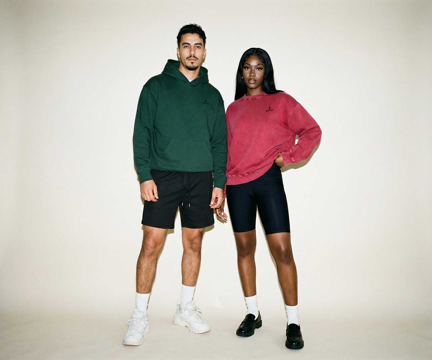Two people wearing green and pink hoodies with black shorts on a plain background