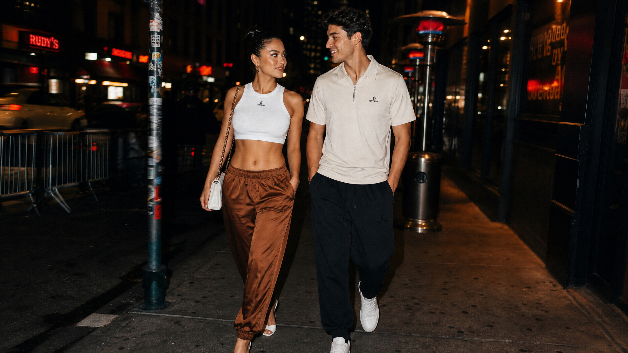 A man and a woman walking hand-in-hand on a city sidewalk at night. The woman wears a white baby tee and brown track pants, while the man wears a white polo shirt, black pants, and white sneakers.