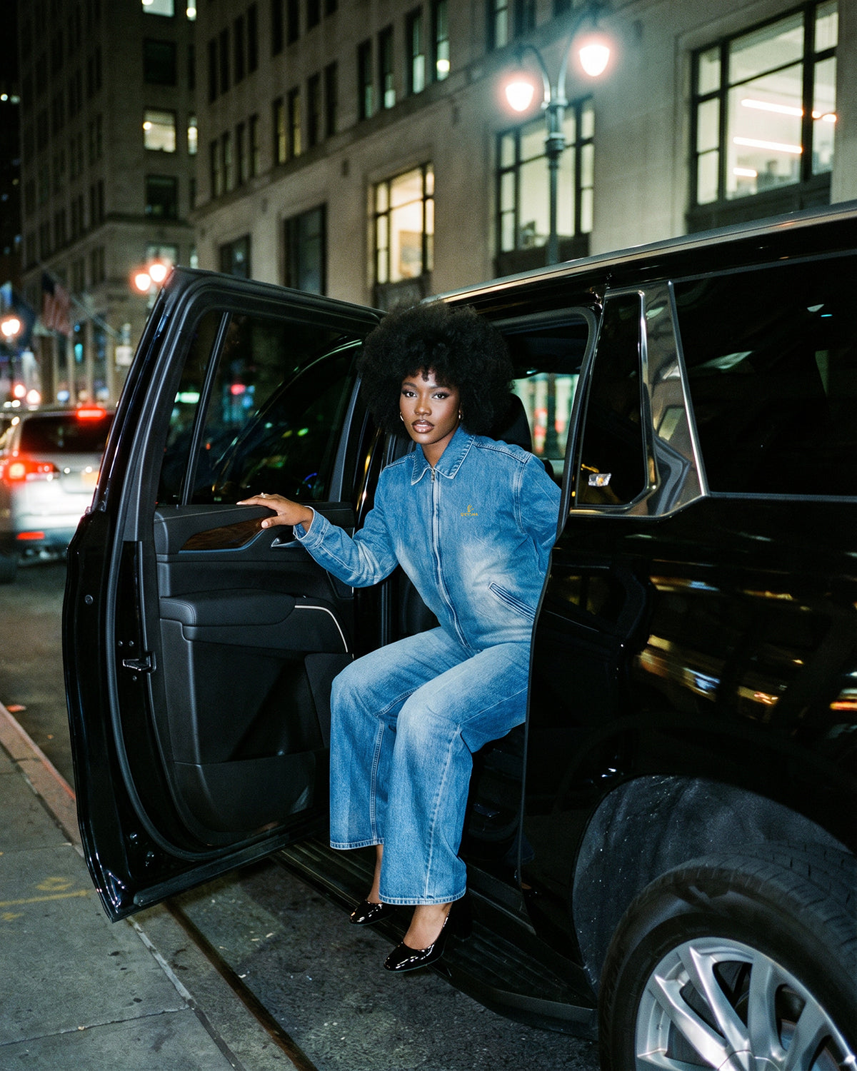 Woman in a blue denim outfit getting out of a black SUV on a city street at night.