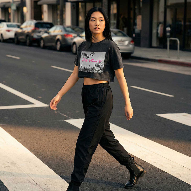 A woman walking across a city street crosswalk wearing a black cropped graphic T-shirt and black pants.