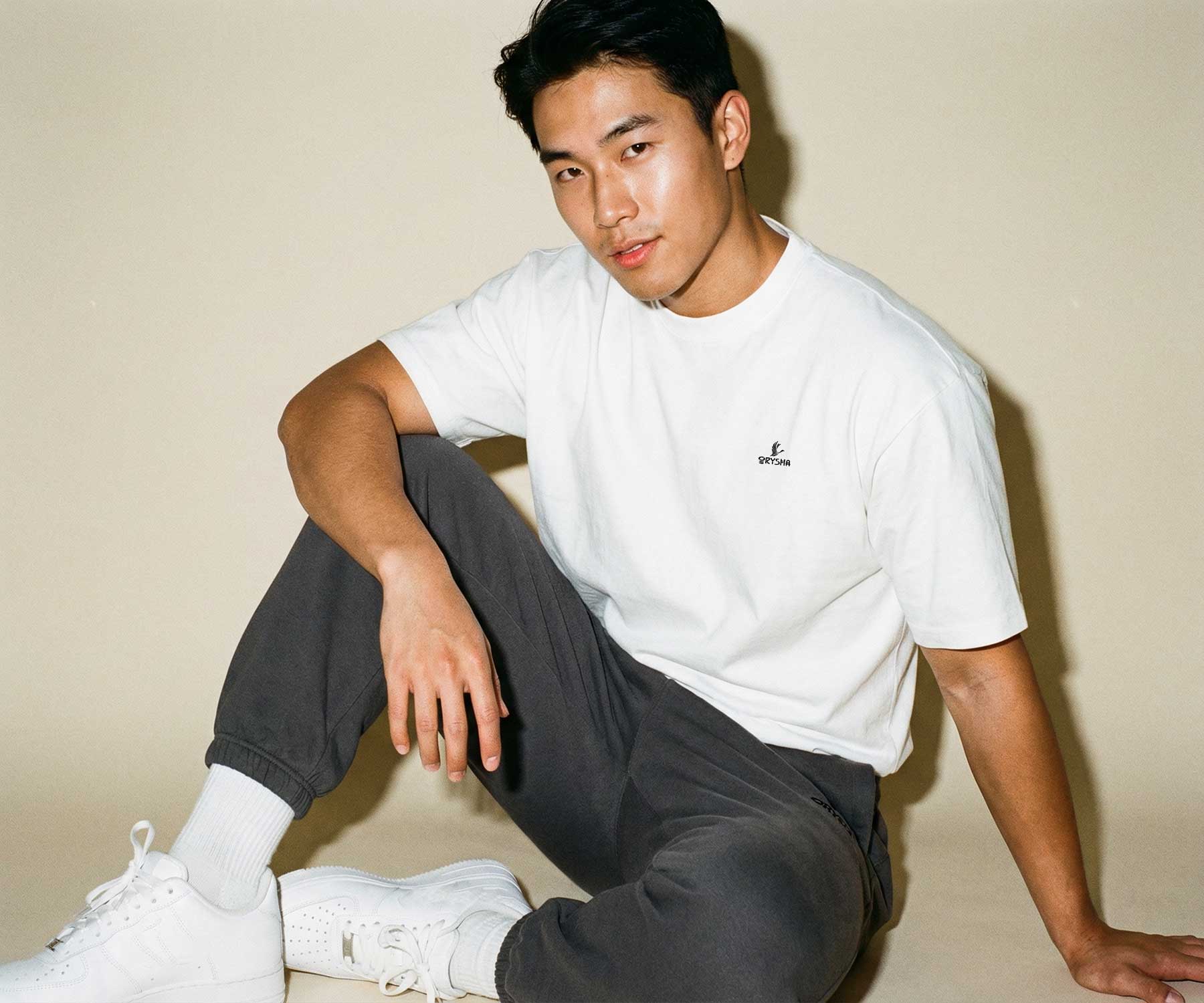 Person wearing a white t-shirt with a logo and dark pants sitting on the floor against a beige wall.